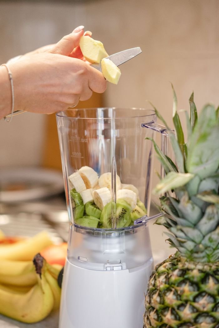 Close-up of hands preparing fresh fruit smoothie with blender, featuring bananas, kiwi, and pineapple.